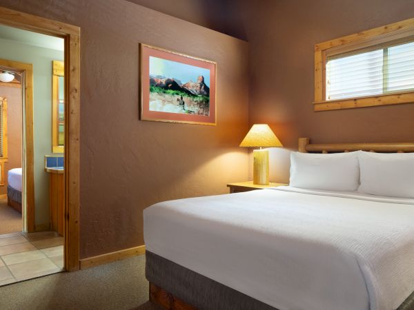 The image shows a cozy bedroom with a large bed, a bedside lamp, a painting above, and a view into another room with a window.