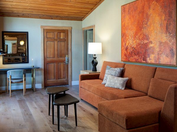 A cozy living room with a brown sectional, a round coffee table, a black lamp, a large abstract orange painting, a wooden console and mirror, and a small desk near a wooden door.