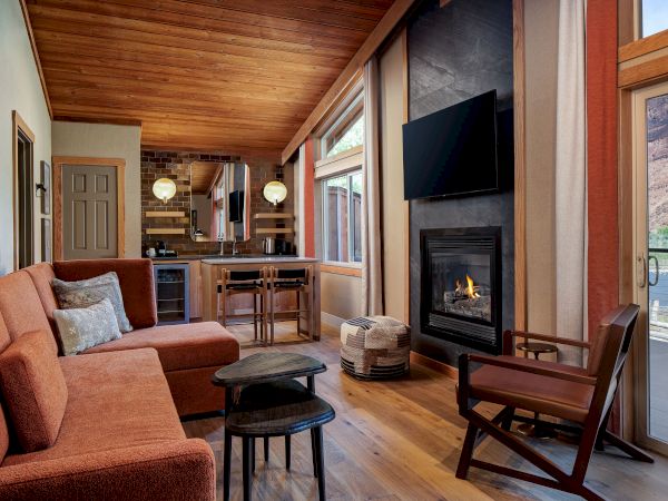 A cozy, modern living room with a wood ceiling, orange sofa, a fireplace, mounted TV, and a seating area near large windows and a glass door.
