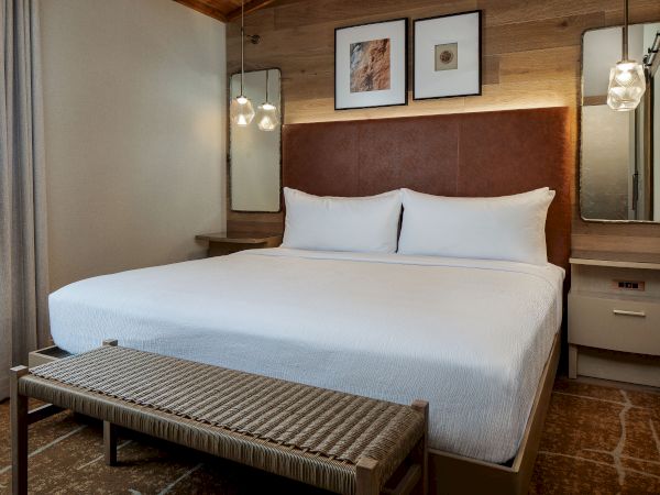 A modern hotel room with a large bed, white linens, bedside tables, wall mirrors, framed art, a wooden headboard, a bench at the foot, and warm lighting.
