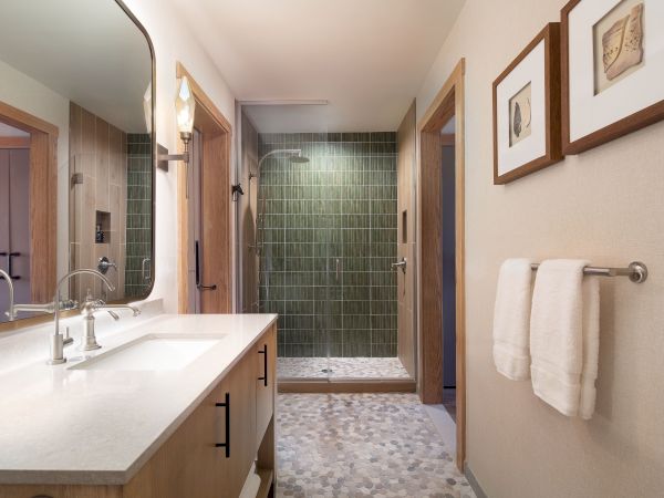 A modern bathroom with a double sink vanity, large mirror, glass shower with green tiles, light wood cabinetry, and framed art above a towel rack.