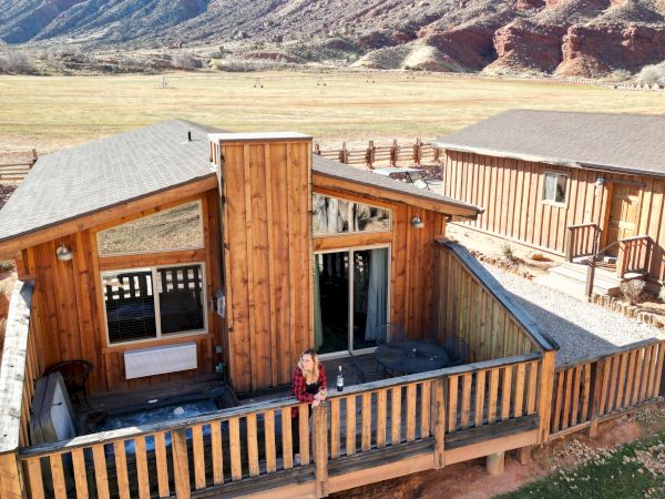 A person stands on the wooden deck of a cabin surrounded by grass, with a backdrop of rocky cliffs and a clear sky.