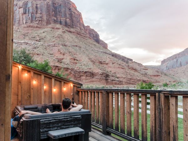 A person relaxes in an outdoor hot tub on a wooden deck, overlooking a scenic canyon landscape with warm string lights.