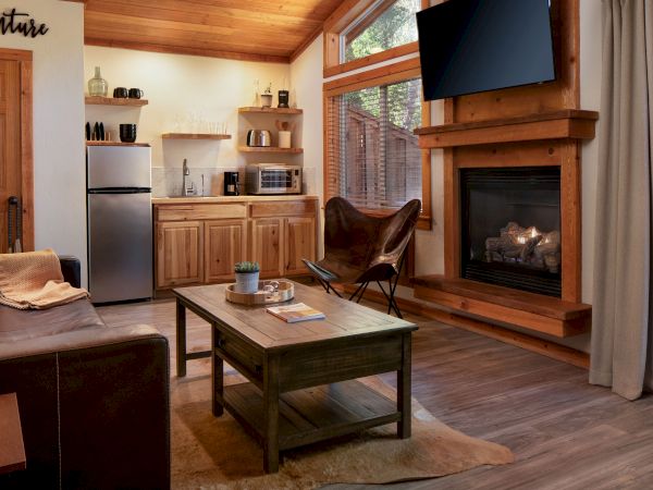 The image shows a cozy living space with a leather couch, wooden furniture, a kitchenette, a fireplace, and a large window.