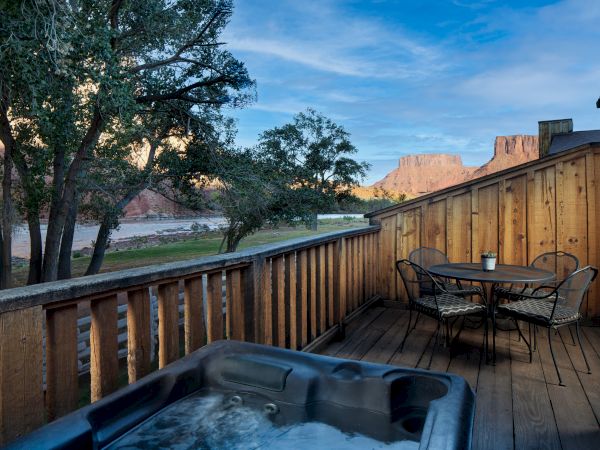 A wooden deck with a hot tub and table set, overlooking trees and distant red rock formations under a blue sky.