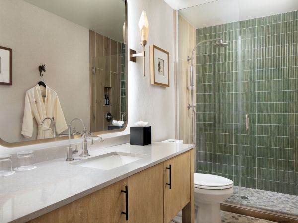 A modern bathroom with a double sink vanity, wooden cabinet, large mirror, wall sconce, robe on a hook, and a green-tiled glass shower enclosure.
