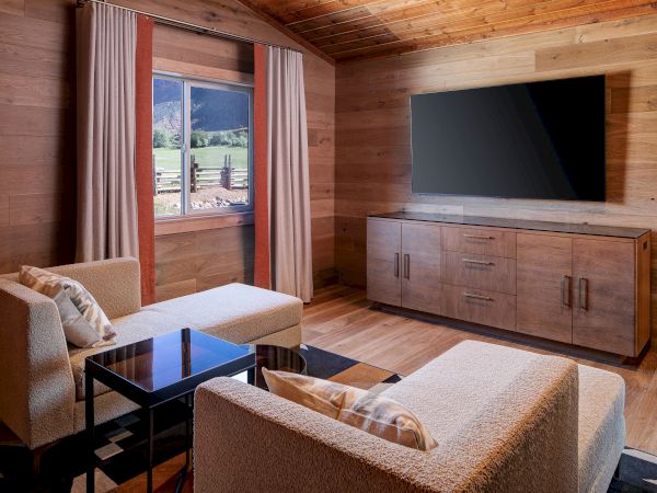 A cozy living room with wood-paneled walls, two beige sofas, a coffee table, a large flat-screen TV, and a wooden cabinet, plus a window with curtains.