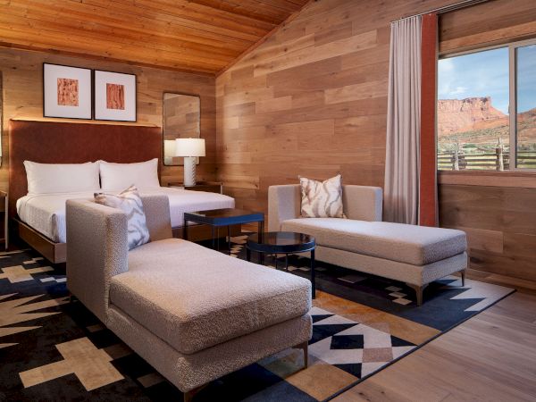 A cozy wood-paneled hotel room with two beds, a seating area, and a large window showing a desert landscape, stylish lamps, and modern decor.