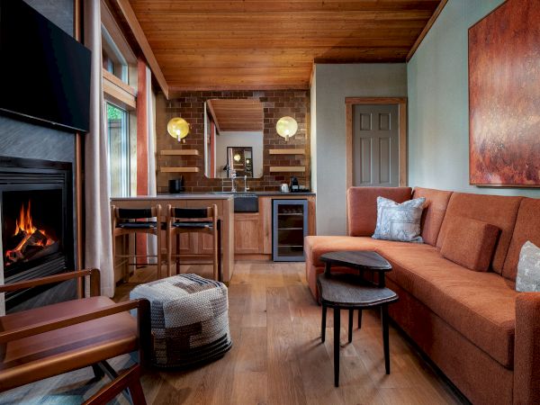 Cozy living room with a fireplace, orange sofa, pendant lights, wood accents, and a small kitchen bar in the background.