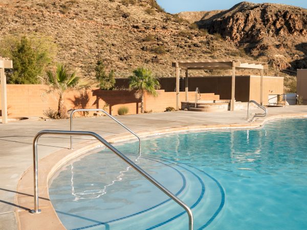 A serene outdoor swimming pool with clear water, surrounded by desert landscapes and mountains under a clear blue sky.