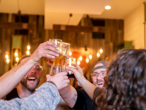 A group of friends toasting with beer in a lively bar, cheering and smiling as they raise clinking glasses to celebrate.