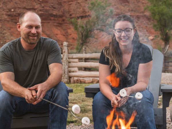 Two adults roasting marshmallows by a campfire, smiling, outdoors with rocks and a wooden fence in the background.