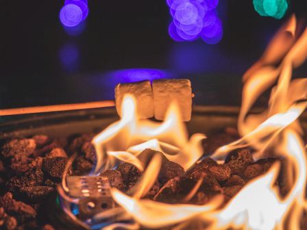 A campfire with glowing flames over rocks, a marshmallow on a stick roasting nearby, and colorful bokeh lights in the blurred background.