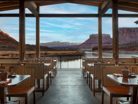 A lakeside dining area with wooden chairs and tables on a covered deck, overlooking a desert canyon and calm water at sunset.