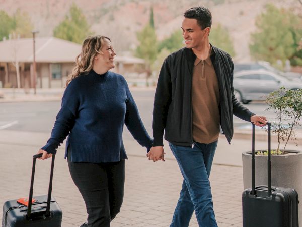 A couple walks hand-in-hand with rolling luggage at a scenic outdoor area, smiling and enjoying their trip together.