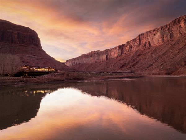 A serene desert canyon at sunset, with a calm river reflecting the pink-orange sky and rugged cliffs, a small building perched along the shore.