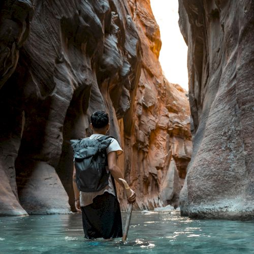 A traveler wades a turquoise river in a canyon with a backpack, holding a paddle as cliffs tower overhead.