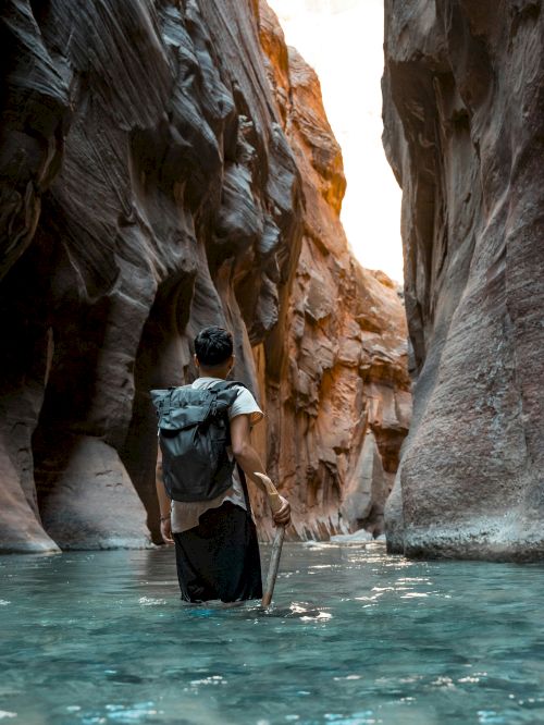 A traveler wades a turquoise river in a canyon with a backpack, holding a paddle as cliffs tower overhead.