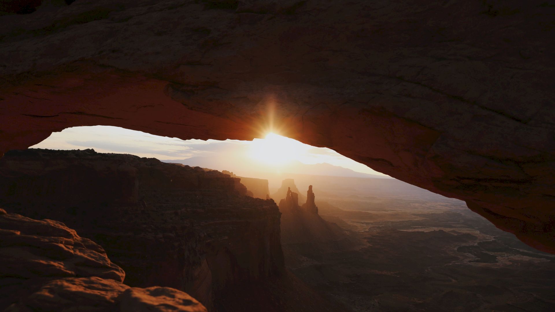 Sunrise pours through a cave opening onto a desert canyon, casting warm light over distant rock formations.