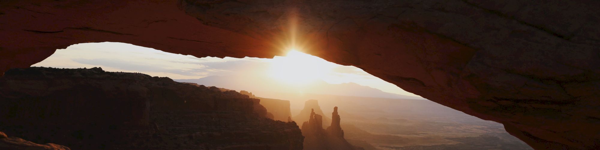 Sunrise pours through a cave opening onto a desert canyon, casting warm light over distant rock formations.